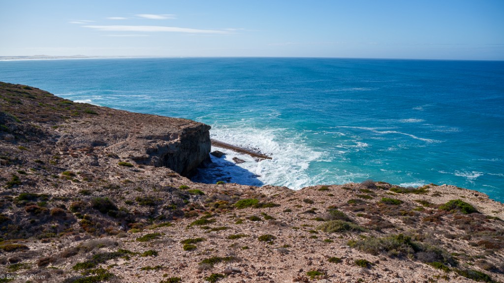A coastal view featuring rugged cliffs and the ocean waves breaking against the shore under a clear blue sky.