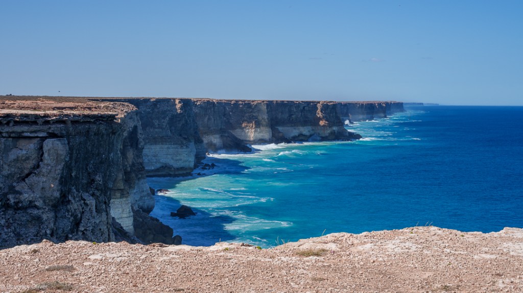 A scenic view of dramatic coastal cliffs overlooking turquoise ocean waters under a clear blue sky.