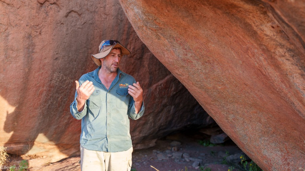 A man stands under a large rock formation, gesturing as he speaks. He wears a blue shirt and a wide-brimmed hat, with sunlight illuminating the scene.