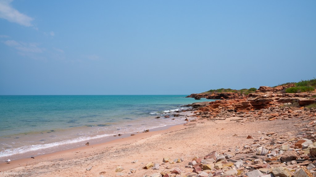 A scenic beach with soft pink sand, rocky cliffs, and calm turquoise waters under a clear blue sky.