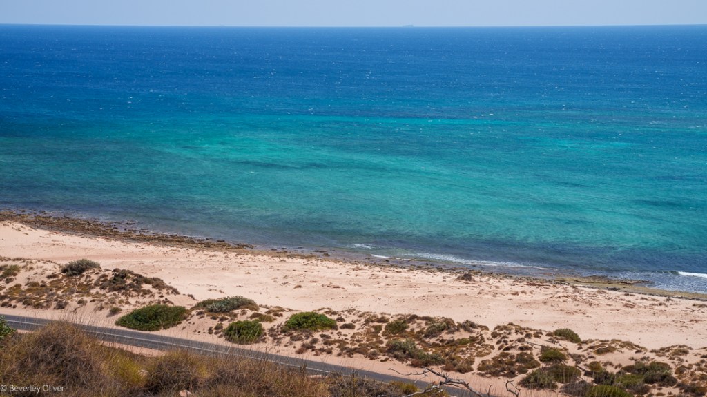 A scenic view of a sandy beach with turquoise waters, featuring gentle waves lapping at the shore and vegetation along the beach.