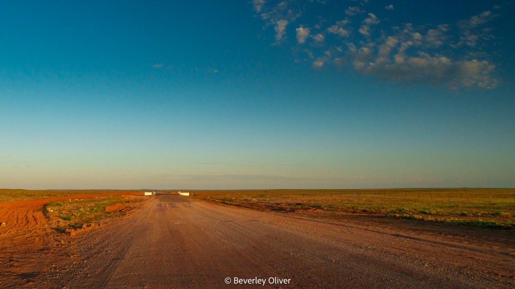 A wide, open dirt road leading towards the horizon, with a clear blue sky and sparse vegetation on either side.