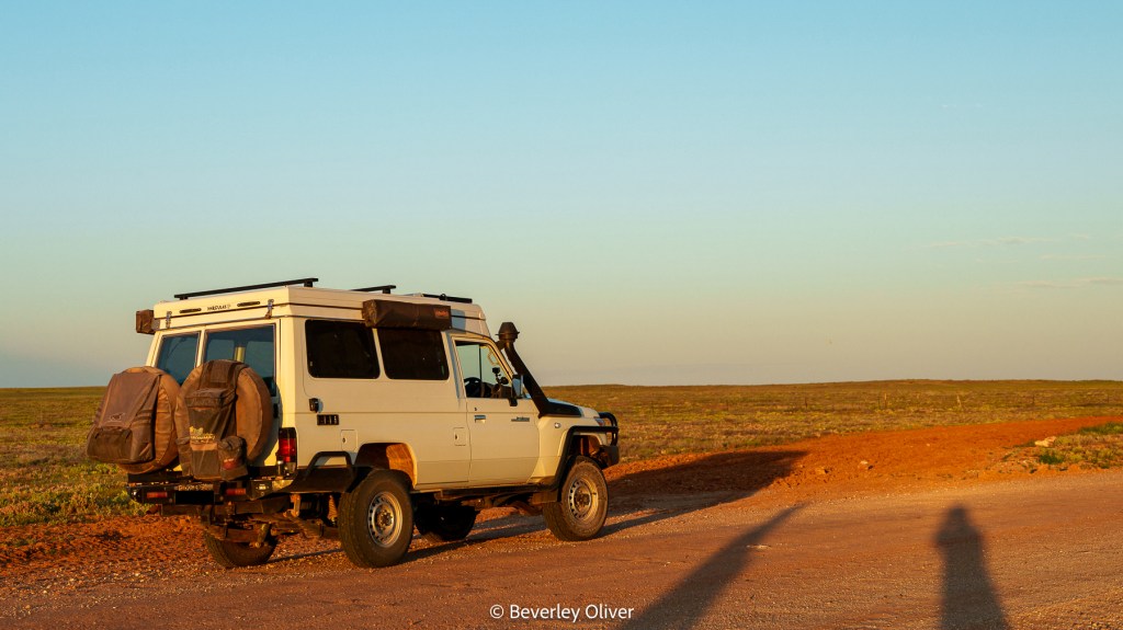 A white 4x4 vehicle parked on a dirt road with camping gear attached, surrounded by an open landscape under a clear blue sky.