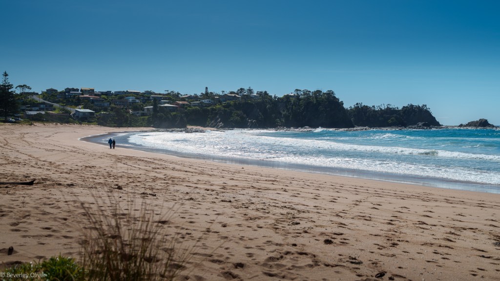 A couple walking hand in hand along a sandy beach with gentle waves under a clear blue sky, overlooking a nearby coastal town.
