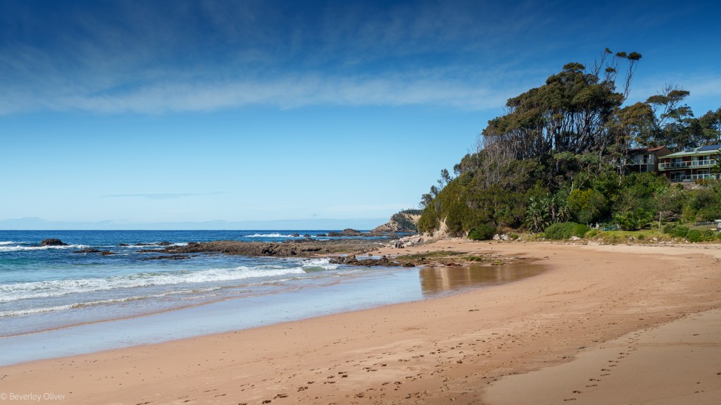 A serene beach scene with gentle waves lapping at the sandy shore, framed by a rocky coastline and lush greenery. A clear blue sky with wispy clouds completes the tranquil atmosphere.