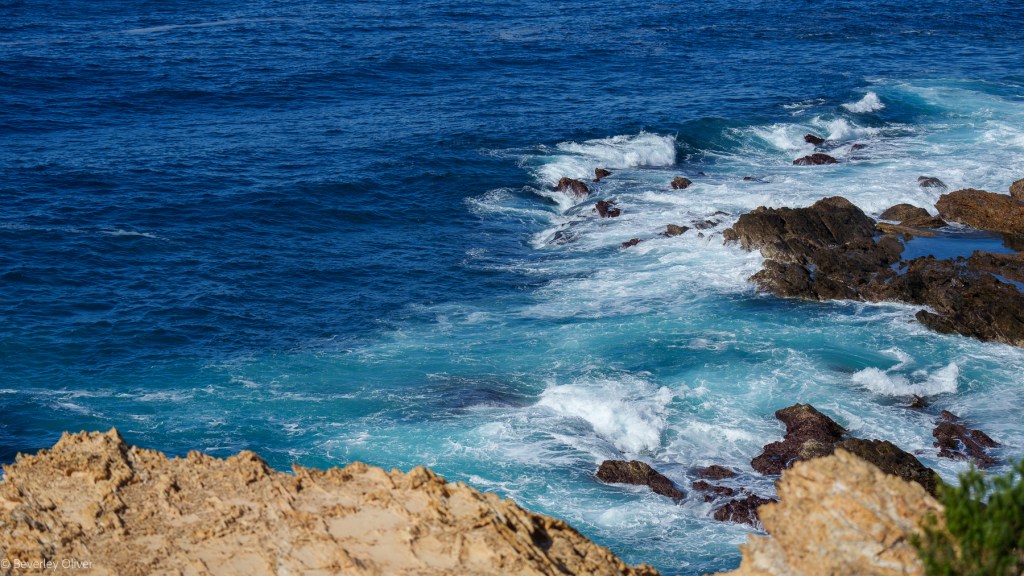 A rocky coastline with waves crashing against the shore, creating foam in the turquoise water.