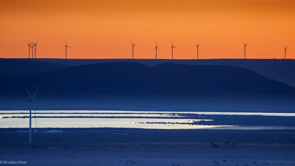Landscape view featuring wind turbines silhouetted against a vibrant orange sunset, with a body of water and rolling hills in the foreground.