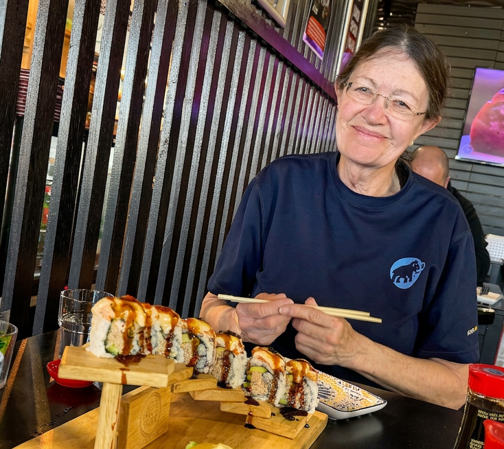 A woman enjoying sushi at a restaurant, holding chopsticks and smiling, with a plate of sushi displayed on a wooden stand.