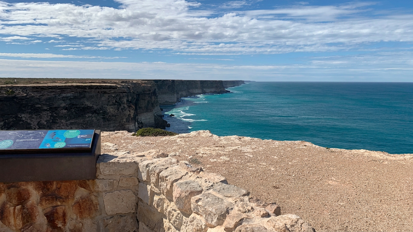 IMG_3508 A scenic coastal view featuring rugged cliffs and vibrant blue ocean waters under a partly cloudy sky. A stone wall and informational display are visible in the foreground.
