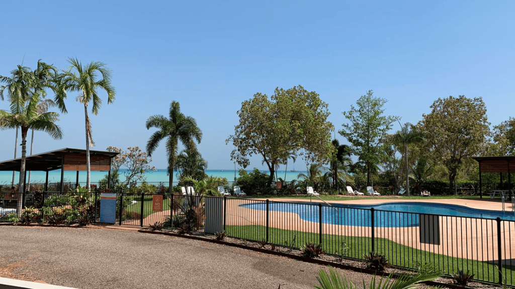 Outdoor swimming pool surrounded by palm trees, with a beach view in the background and clear blue sky.