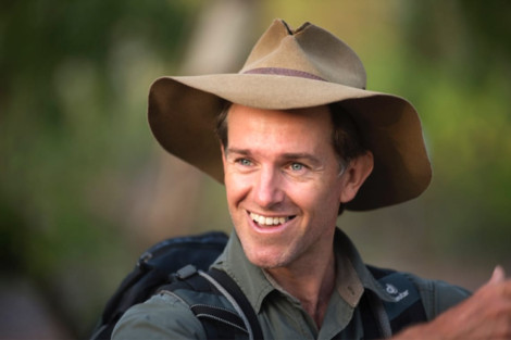 A smiling man wearing a wide-brimmed hat and a backpack, standing outdoors in a natural setting.