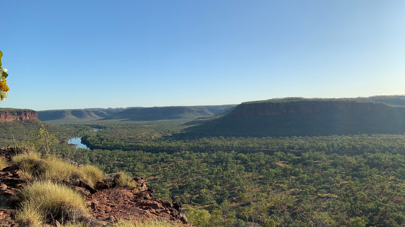 A panoramic view of a lush green valley surrounded by rocky cliffs under a clear blue sky, with a river flowing through the landscape.