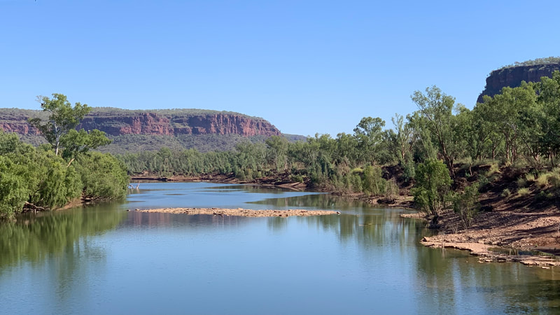A serene river landscape featuring calm waters reflecting the blue sky, surrounded by lush greenery and reddish cliffs in the background.