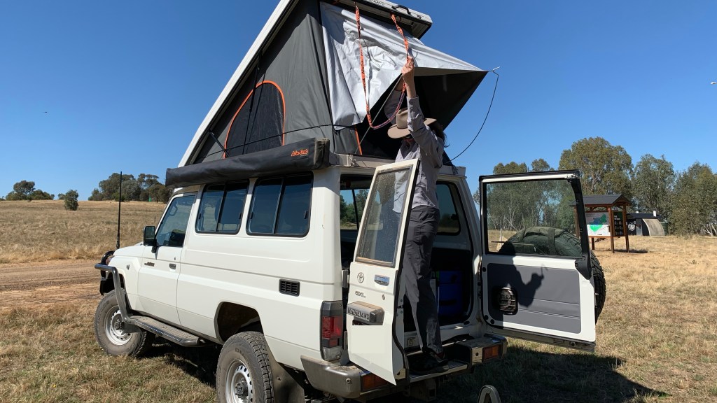 A person is raising the rooftop tent on a white utility vehicle in a grassy outdoor setting under a clear blue sky.