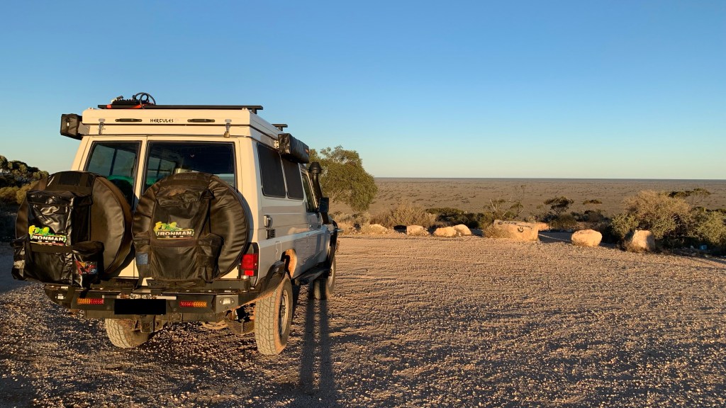 A rear view of a four-wheel drive vehicle parked on a dirt surface with a vast, open landscape in the background under a clear blue sky.