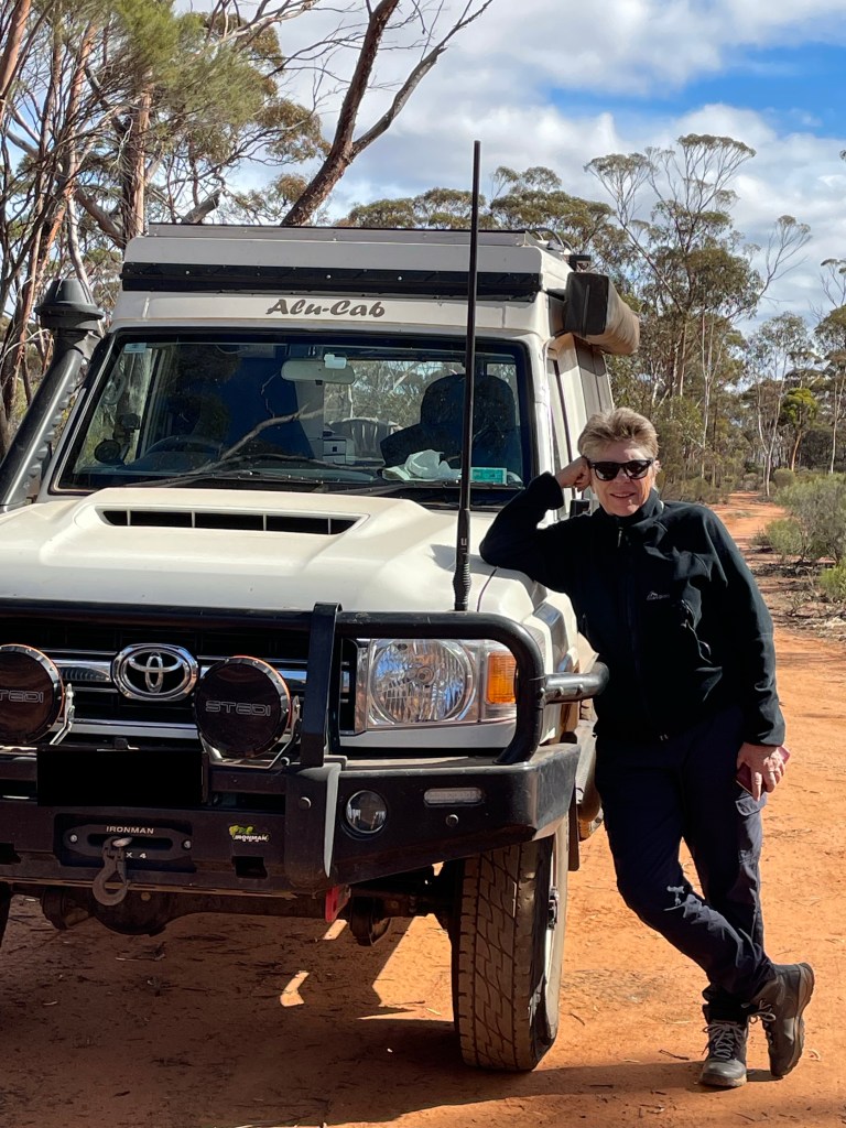 A person leaning against a white Toyota 4x4 vehicle with a rooftop tent, surrounded by a natural landscape of trees and dirt.