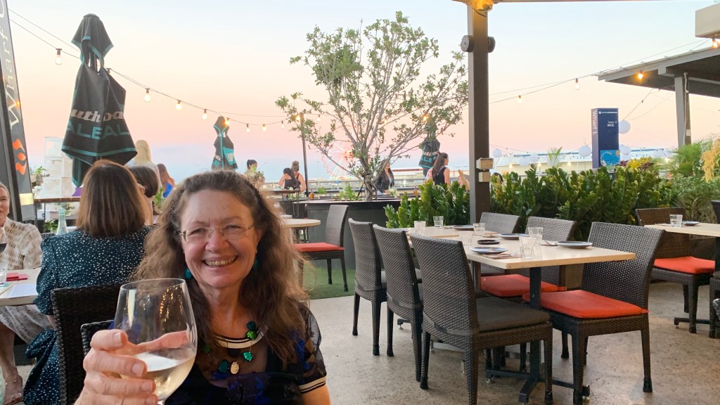 A smiling woman holding a glass of wine at a restaurant table, with outdoor seating and a sunset backdrop featuring string lights and greenery.