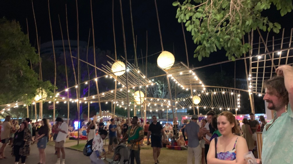 A lively outdoor nighttime festival scene featuring people mingling under decorative lighting and lanterns, with a backdrop of bamboo structures and greenery.