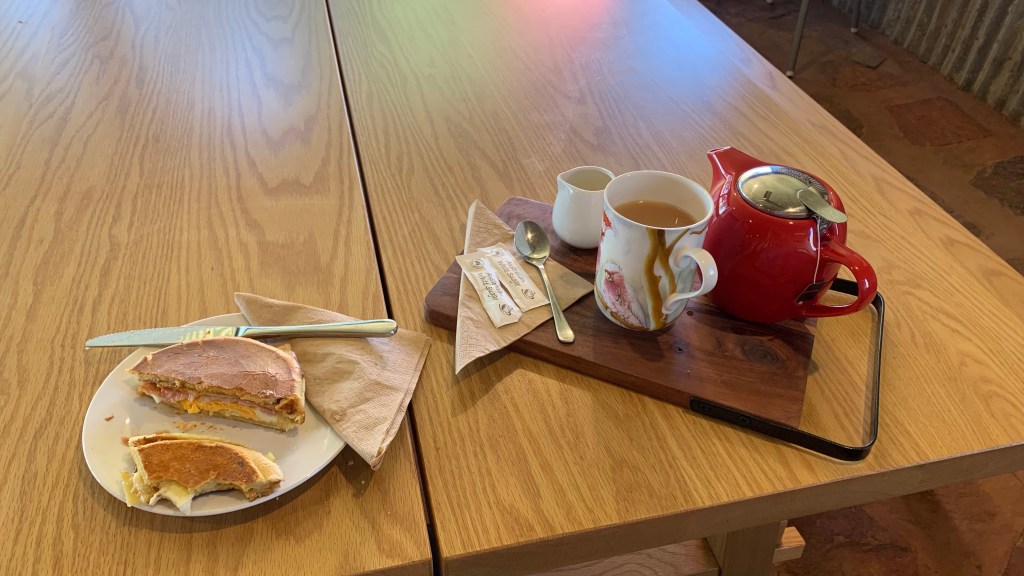 A plate with a slice of sandwich, a knife, and a napkin beside it, next to a wooden tray holding a red teapot, a tea cup with a cream pitcher, and a spoon.