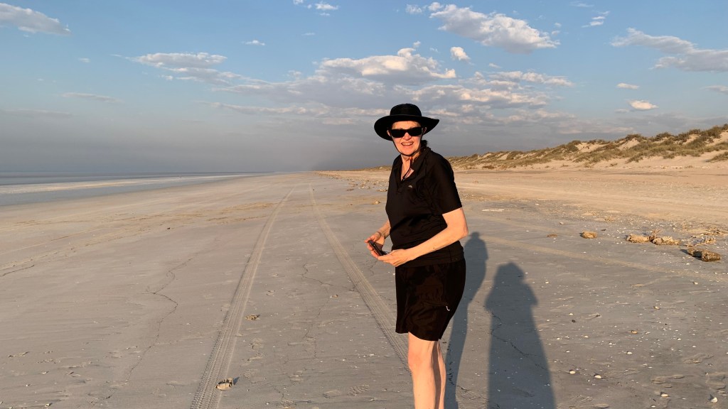 Person standing on a sandy beach wearing a black outfit and hat, with tire tracks in the sand and a blue sky in the background.