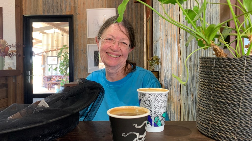 A smiling woman sitting at a table with two coffee cups and a decorative plant in the foreground, surrounded by a rustic interior.