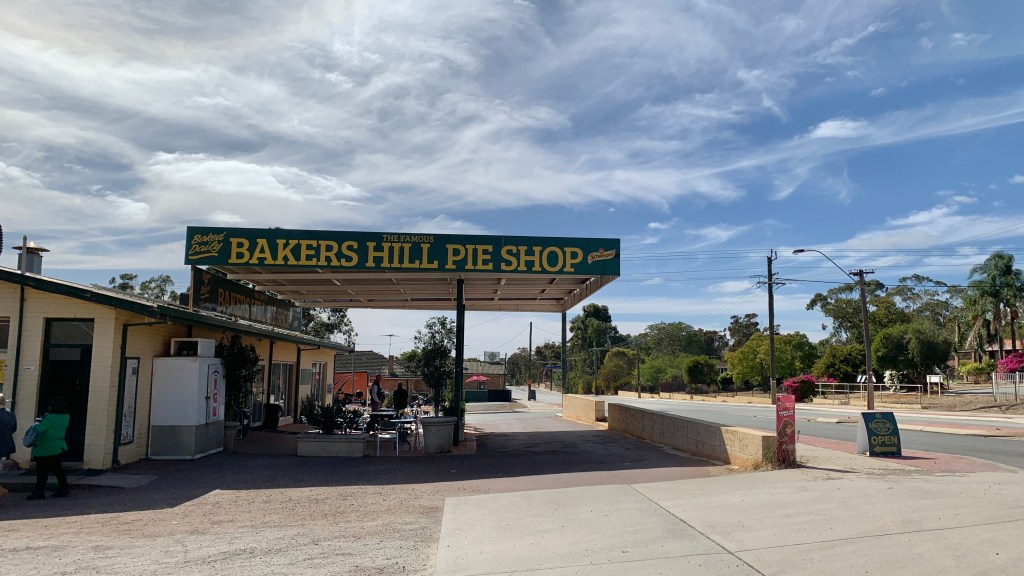 Exterior view of Bakers Hill Pie Shop with a green sign, outdoor seating area, and clear blue sky.