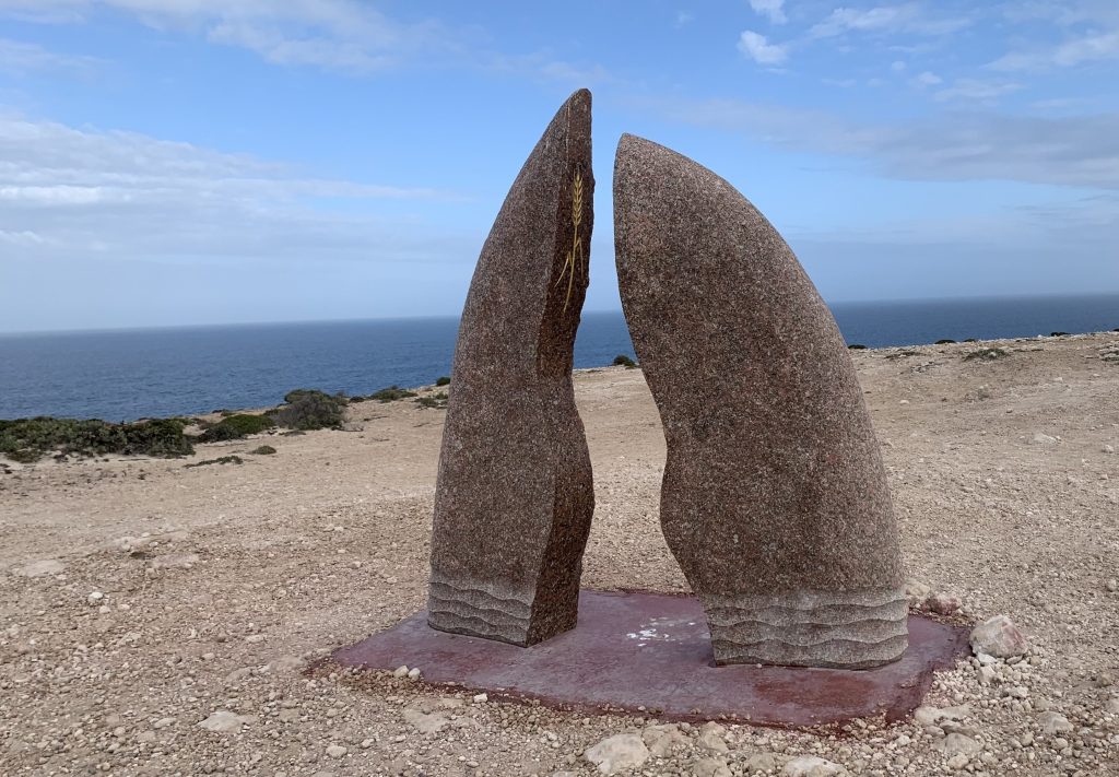 A large stone sculpture with two curved, pointed halves standing on a rocky landscape, overlooking the ocean under a partly cloudy sky.