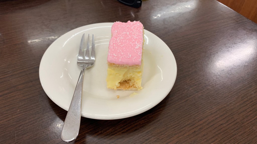 A slice of layered cake with a pink frosting on top, served on a white plate with a fork.