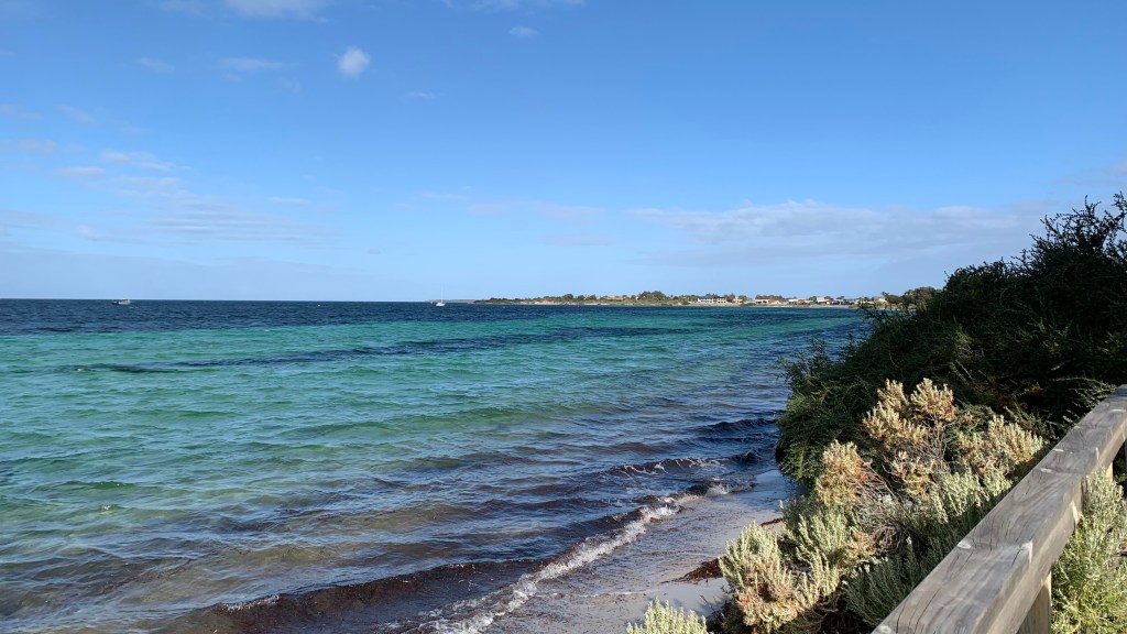 A tranquil coastal scene showing clear blue-green waters with gentle waves, a sandy beach lined with shrubs, and a distant shoreline under a bright sky.