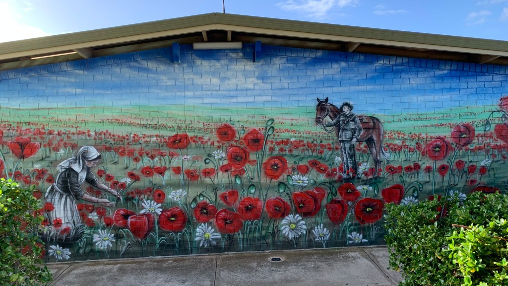 A mural depicting a woman picking poppies in a field, with a soldier holding a horse nearby, set against a blue sky.