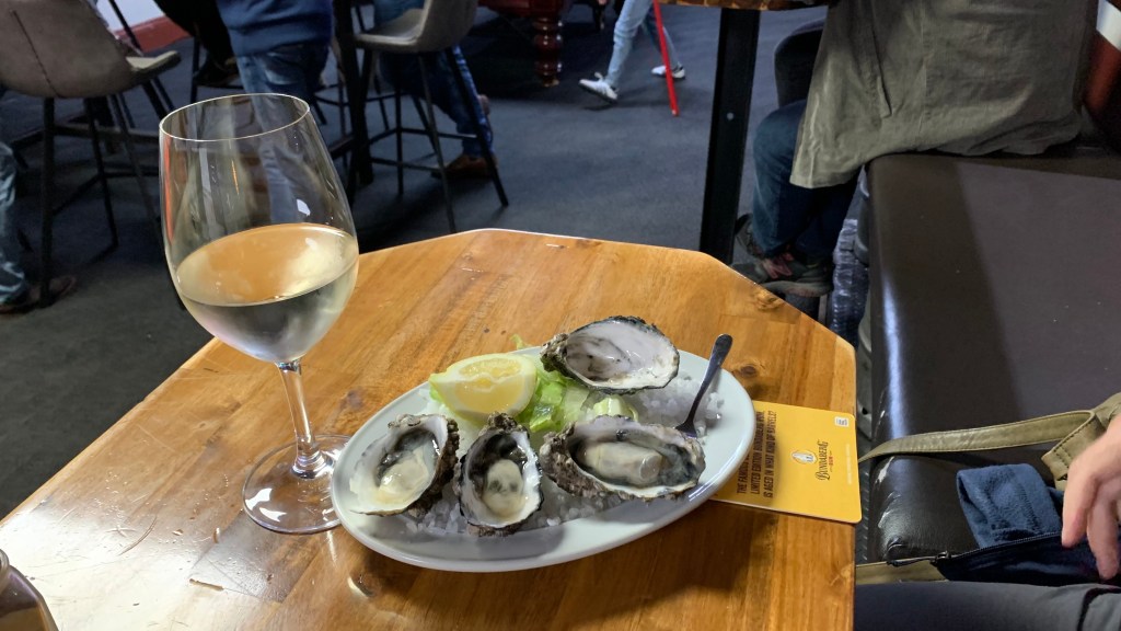 A plate of oysters on ice with a slice of lemon, accompanied by a glass of white wine, on a wooden table in a casual dining setting.