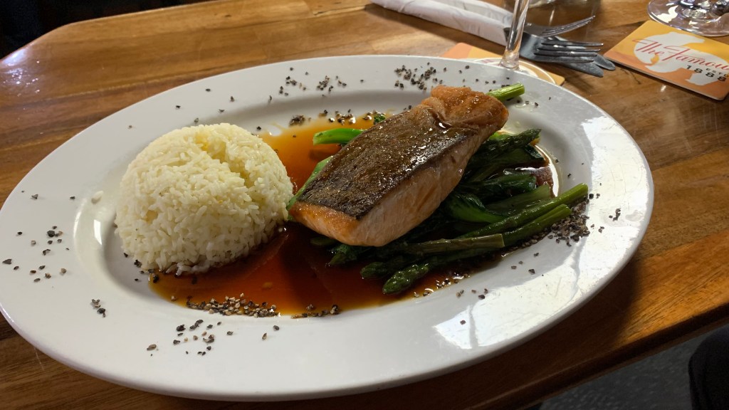 A plate of seared salmon fillet served on a bed of sautéed asparagus, complemented by a side of fluffy white rice and drizzled with a savory sauce.