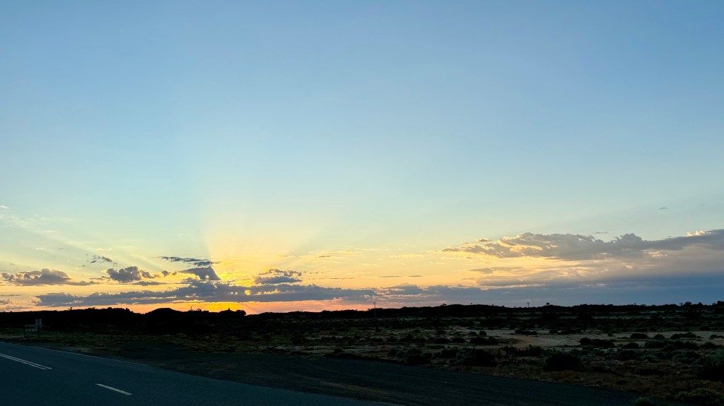 Scenic view of a sunset over a vast landscape, with rays of light emerging from behind clouds, and a quiet road in the foreground.