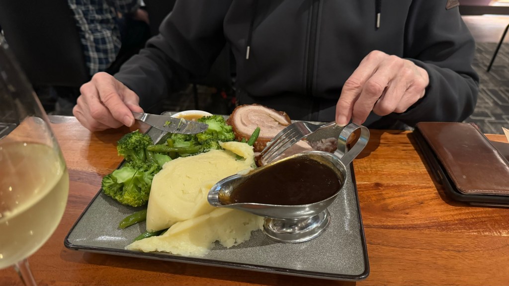 A person using a fork and knife to enjoy a plated meal with mashed potatoes, broccoli, green beans, and sliced meat, with a small boat of gravy on the side.