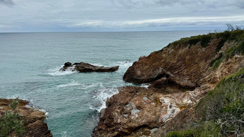 A rocky coastline with waves crashing against the stones, under a cloudy sky.