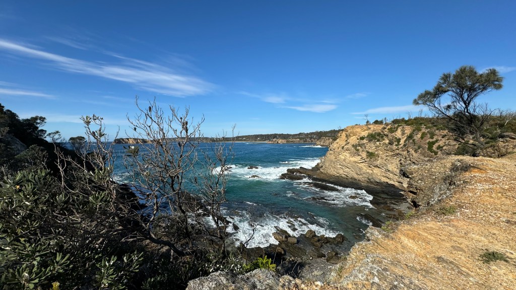 Scenic coastal view featuring rocky cliffs and turquoise waves crashing against the shore, under a clear blue sky.