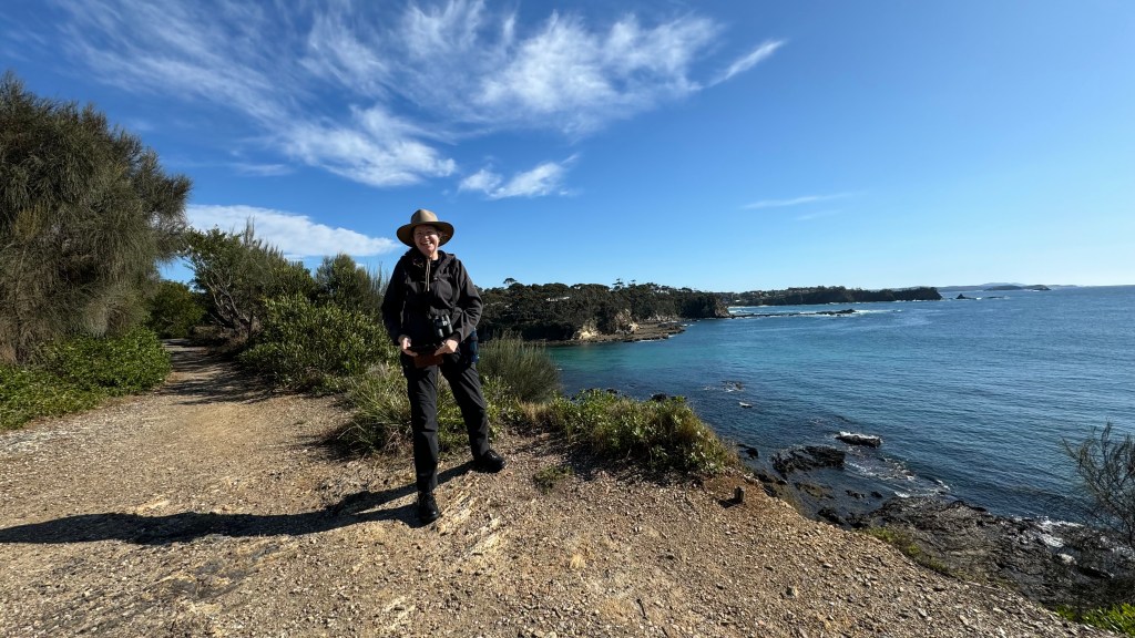 A person standing on a coastal path with a camera, wearing a hat, overlooking the ocean under a clear blue sky with some clouds.