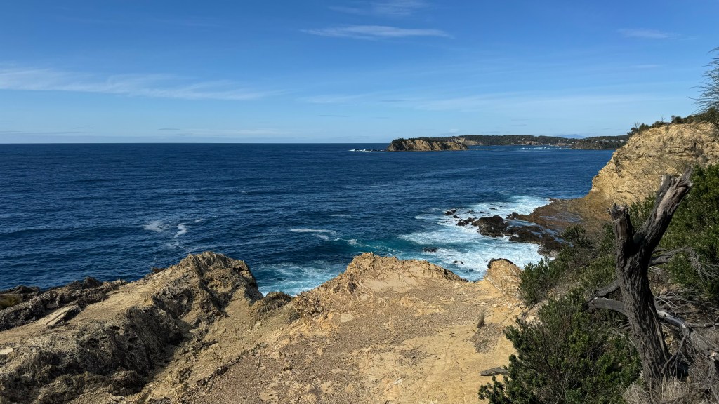 Coastal landscape with rocky cliffs overlooking the ocean and distant hills under a clear blue sky.