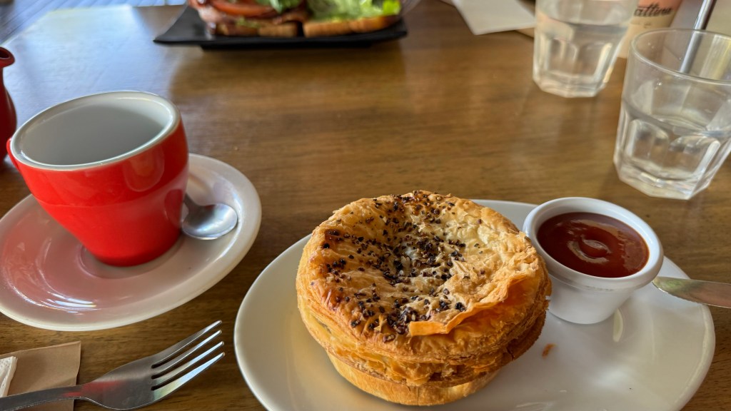 A red coffee cup on a saucer beside a flaky pastry topped with black seeds, served with a small dish of ketchup, on a white plate.