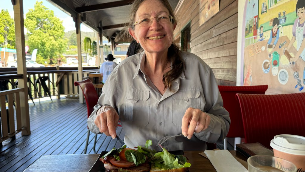 A smiling woman enjoys a meal at a restaurant, holding a fork and a knife with a plate of salad and meat in front of her.