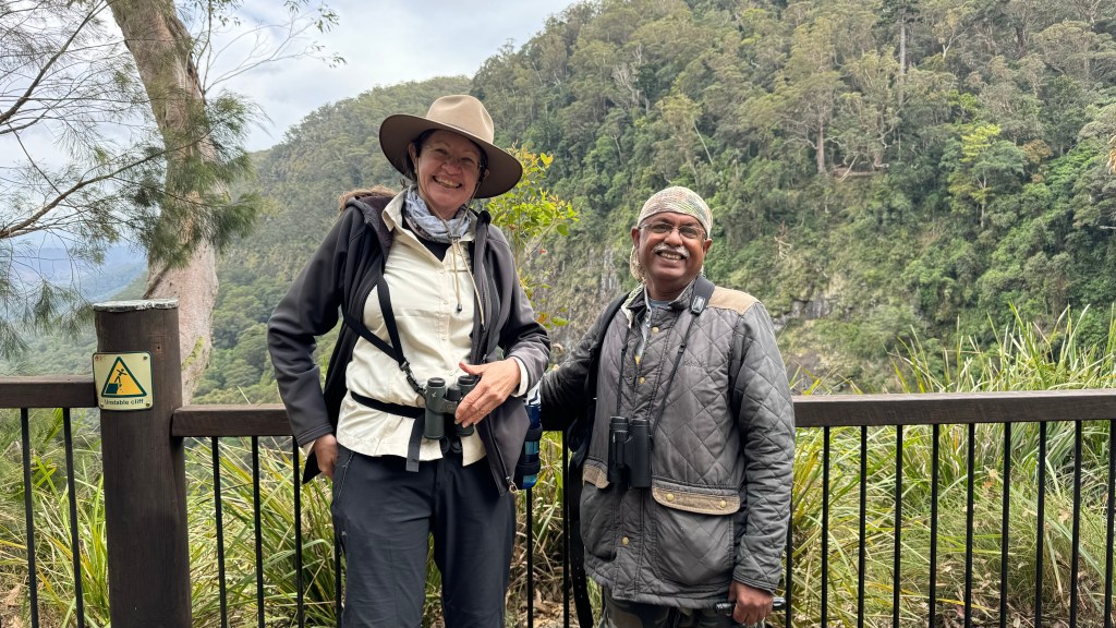 Two hikers posing for a photo at a scenic viewpoint, surrounded by lush greenery and a steep cliff, with a warning sign visible in the background.