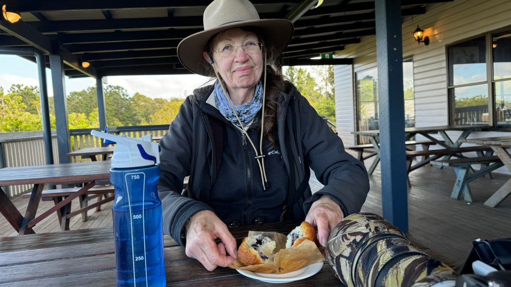 A woman sitting at a table outdoors, smiling while holding a piece of food on a plate. A blue water bottle is on the table next to her, with picnic tables and greenery in the background.