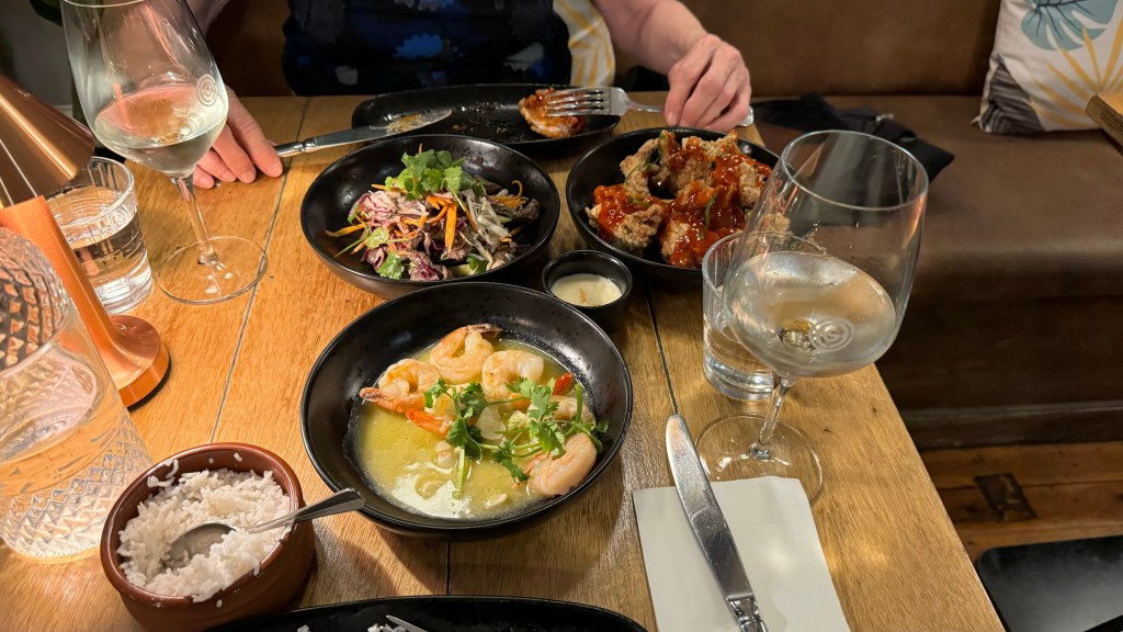 A table set with various dishes including shrimp in a broth, a colorful salad, and a dish of fried food with sauce. There are two glasses of wine, a bowl of rice, and a water glass. A hand holding a fork is also visible.