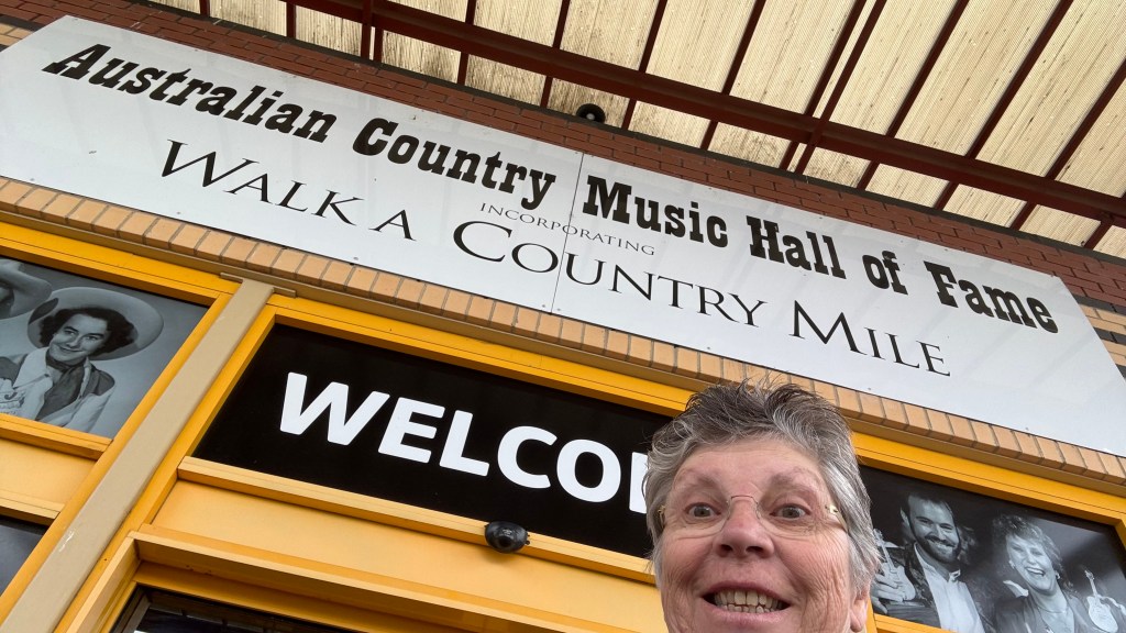 A person smiling in front of the Australian Country Music Hall of Fame entrance sign, which features the text 'WELCOME' and 'Incorporating Walk A Country Mile'.