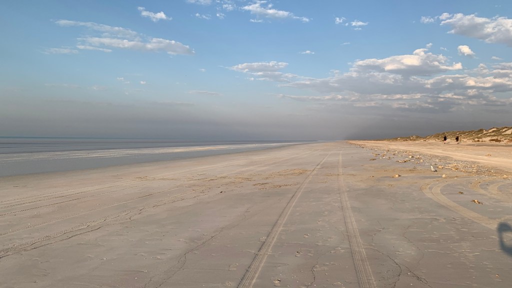A tranquil beach scene with tire tracks leading along the sandy shore under a partly cloudy sky.