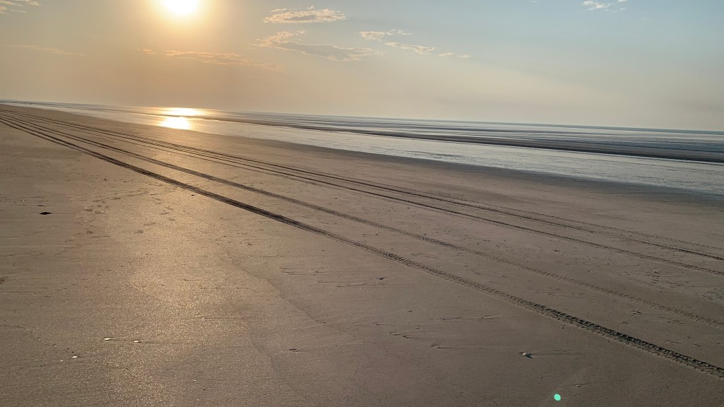 A serene beach scene at sunset with tire tracks on wet sand, reflecting the sun's warm glow.