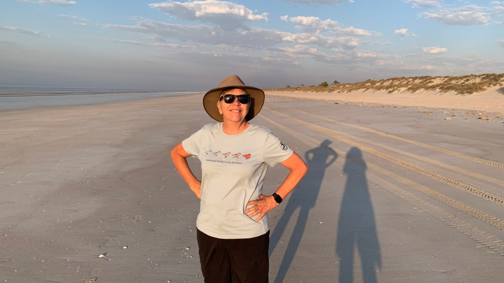A person standing on a sandy beach, wearing a wide-brimmed hat and sunglasses, with hands on hips and a relaxed smile, framed by a scenic shoreline and soft clouds in the sky.