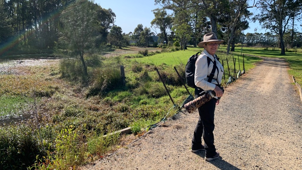 A person standing on a gravel path beside a grassy area and a small water body, wearing a hat and a backpack, with a camera bag slung over their shoulder.